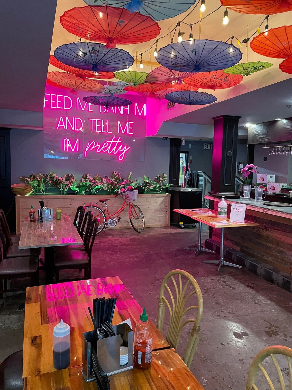Dining room under painted paper parasols, with the pink neon sign reading Feed me banh mi and tell me I'm pretty